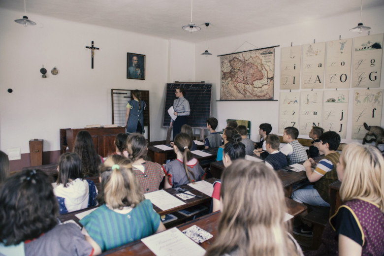 A group of children sit in a reconstructed historical classroom while the teacher leads a math lesson beside a blackboard, surrounded by old maps and teaching charts.