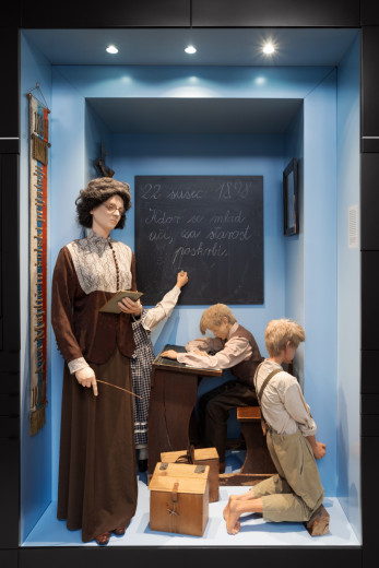 Museum reconstruction of a classroom scene from around 1900: a teacher with a pointer, a pupil writing on the blackboard, another at a desk, and a punished boy kneeling in the corner.