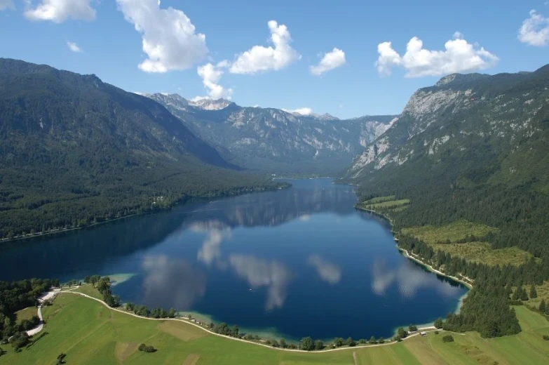 Aerial view of lake Bohinj, surrounded by hills on both sides of the river, and a huge grass field in front of the lake. 
