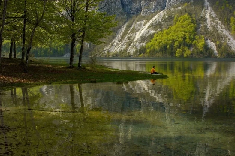 A boy in distance, sitting at the edge of a bank, looking at the lake. 