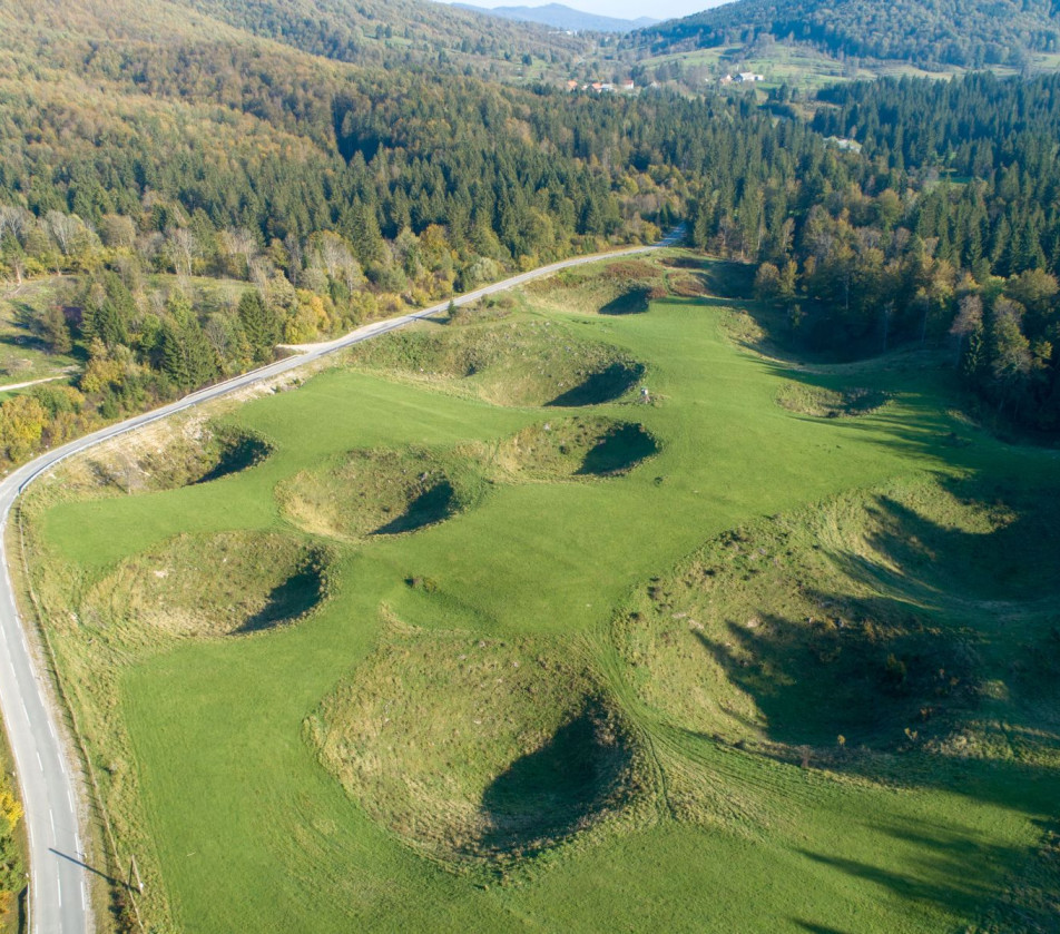 An aerial view of a wide surface of grass with a lot of sinkholes, next to a road and forests. 