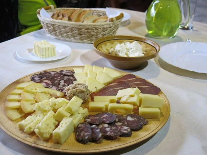 A table with a wooden plate with cheese and sausage, a small basket with bread in the background. 