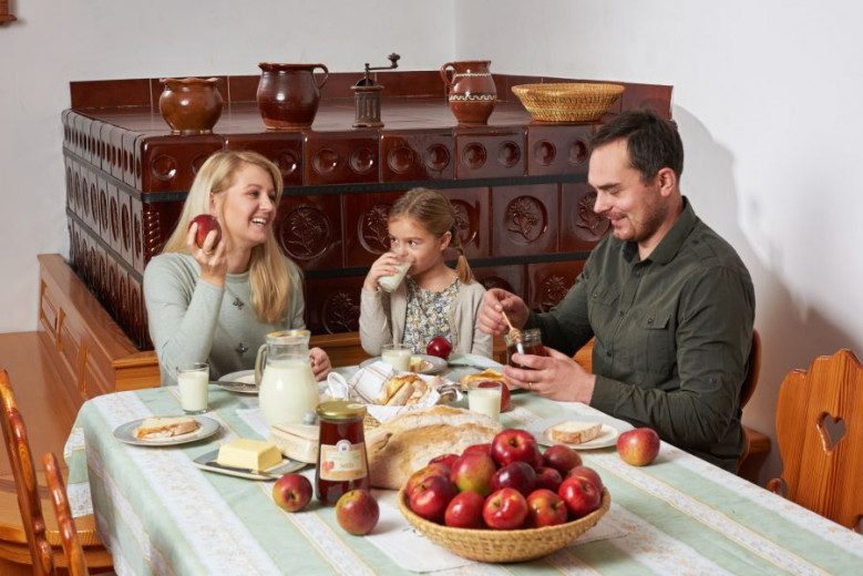 Mama, oče in hči sedijo za mizo in zajtrkujejo. Na miziso kruh, mleko, maslo, med in jabolka.