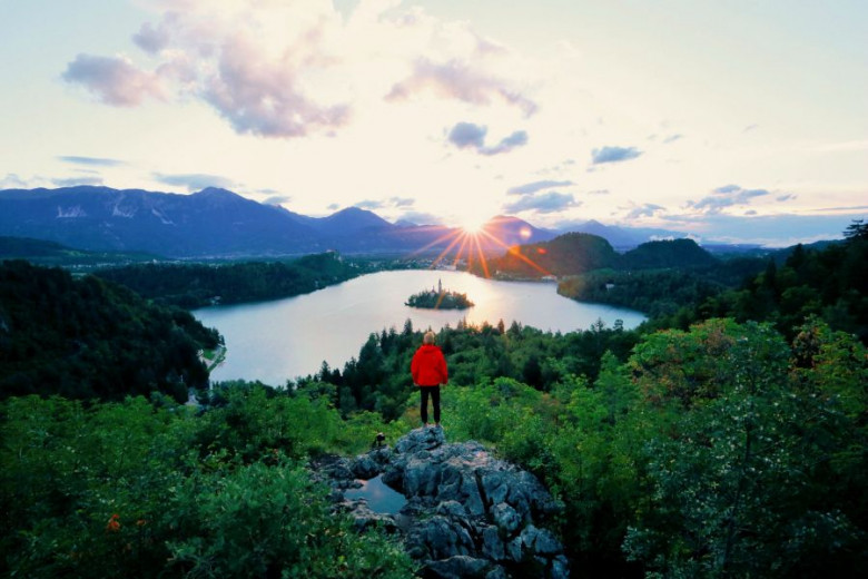A hiker stands on a rock and looks towards Lake Bled. The sun rises behind the hills.