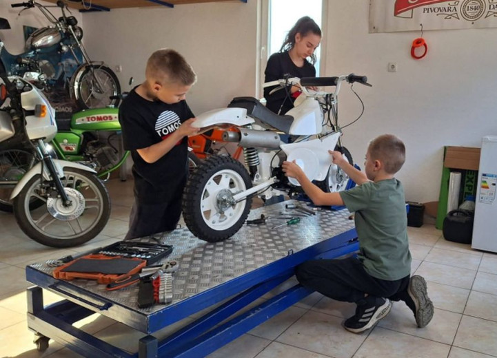 Three children are joyfully working on a small motorbike in a garage. Tools are scattered on the platform. Several motorcycles are visible in the background.