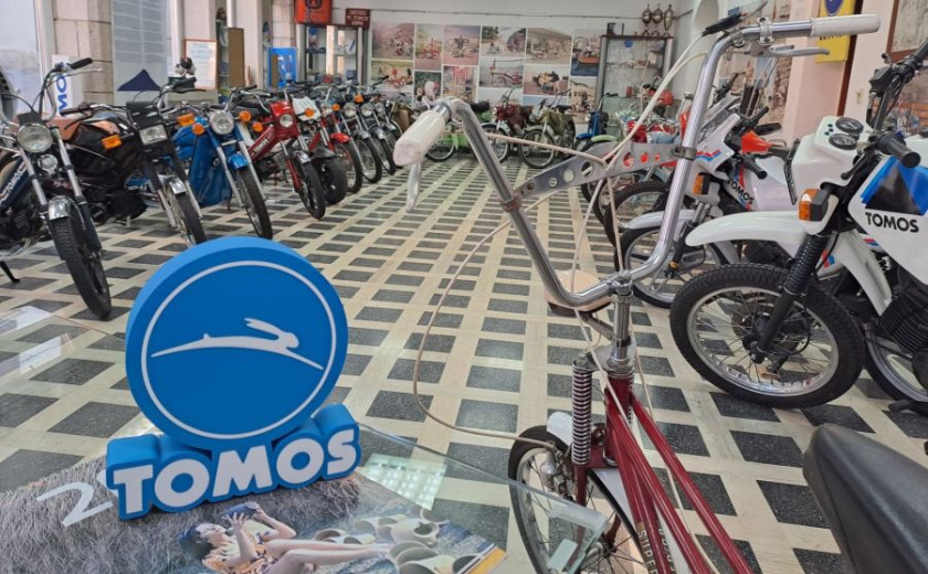 Vintage motorcycle showroom with various retro bikes lined up on a checkered floor. A blue "Tomos" sign in the foreground adds a nostalgic touch.