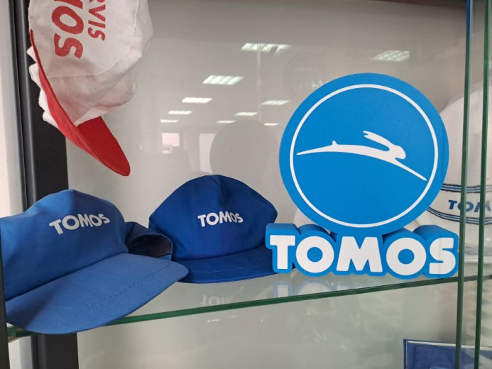 Display shelf with blue "TOMOS" caps and a logo featuring a leaping rabbit. The logo is blue and white with a playful, sporty tone.