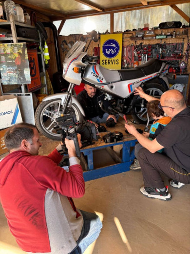 A vintage motorcycle is being repaired by three people in a workshop filled with tools. A cameraman captures the scene, creating a collaborative and industrious atmosphere.