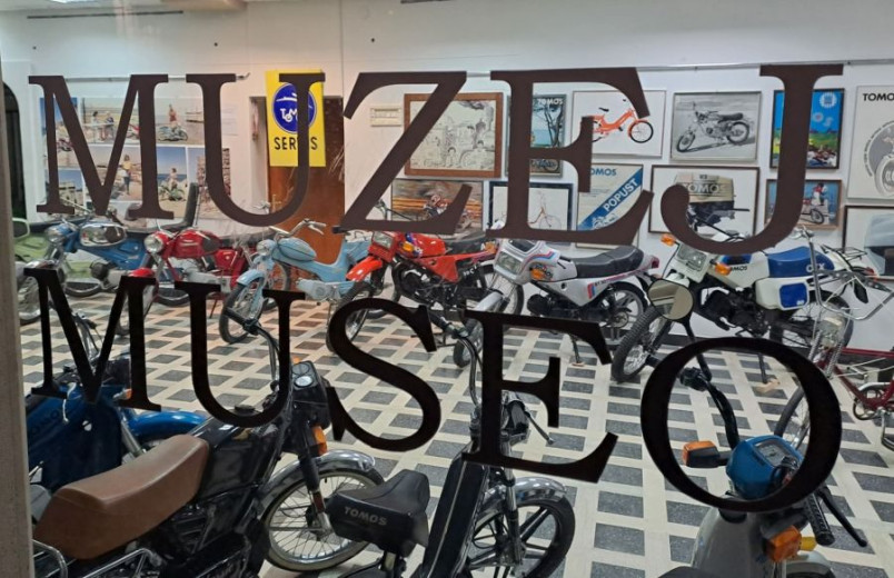 View through a glass window reading "MUZEJ MUSEO" into a vintage motorcycle exhibit, showcasing various colorful bikes on a checkered floor. Nostalgic ambiance.