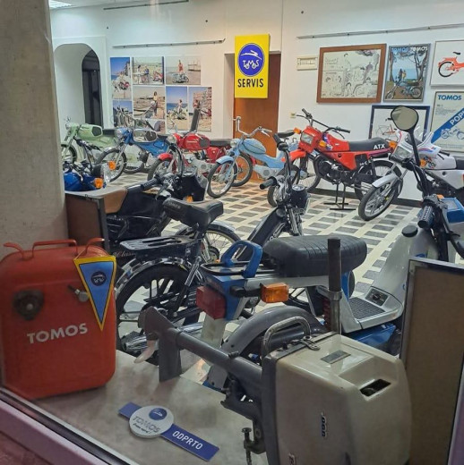 A showroom featuring vintage Tomos mopeds in various colors, arranged on a checkered floor. A wall displays nostalgic photos and a "Tomos Servis" sign. The mood is retro and nostalgic.