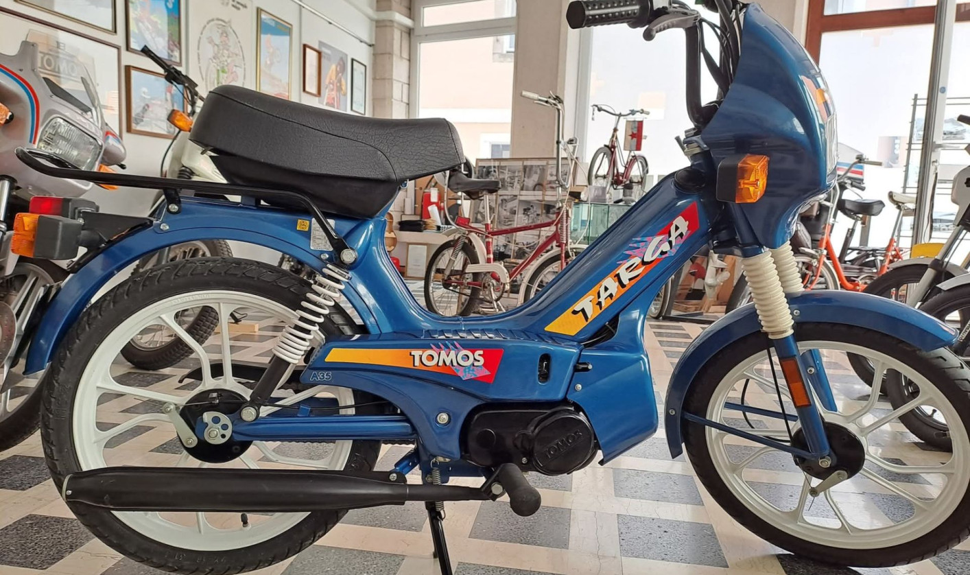 Blue Tomos moped with colorful decals displayed in a showroom with large windows and various vintage bikes. Retro and nostalgic atmosphere.
