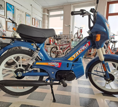 Blue Tomos moped with colorful decals displayed in a showroom with large windows and various vintage bikes. Retro and nostalgic atmosphere.