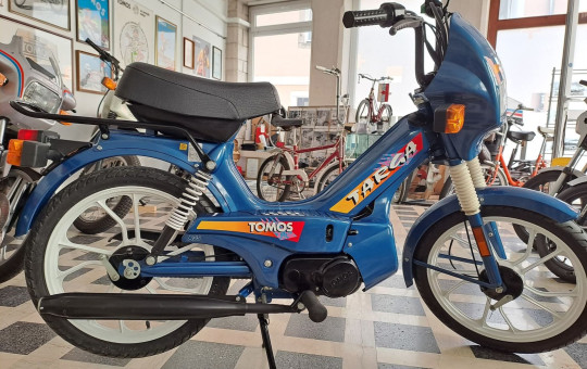 Blue Tomos moped with colorful decals displayed in a showroom with large windows and various vintage bikes. Retro and nostalgic atmosphere.