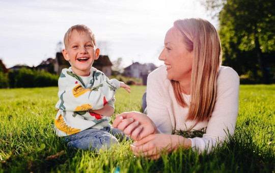 Deček Urban sedi na travi, poleg njega je njegova mama Špela Miroševič, ki ga gleda in se mu nasmiha.