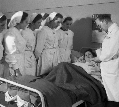 A group of nurses and a doctor stand beside a hospital bed, observing a female patient who is lying under blankets. The doctor appears to be speaking or explaining something to the nurses.
