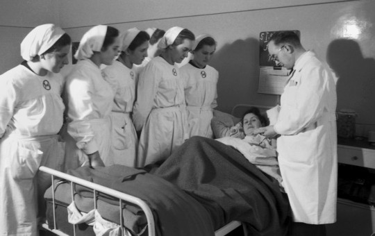 A group of nurses and a doctor stand beside a hospital bed, observing a female patient who is lying under blankets. The doctor appears to be speaking or explaining something to the nurses.