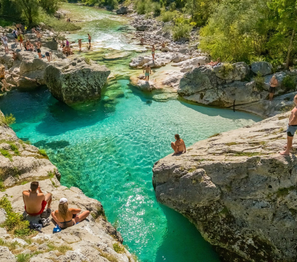People in swimsuits stand on the rocks surrounding one of the Soča River’s natural pools, filled with crystal-clear emerald water.