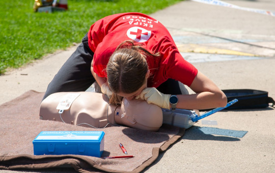 Red Cross volunteer practices CPR rescue breaths on a mannequin outdoors with first aid kit during emergency training.