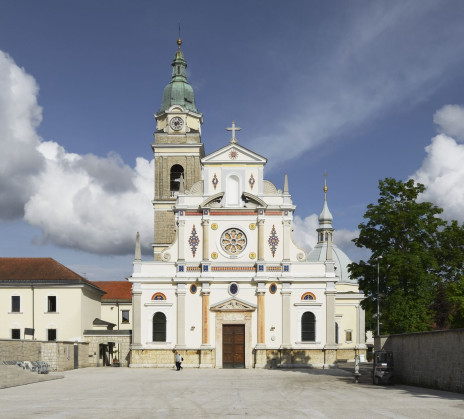 This photo shows the front view of a large church with an ornate white façade, decorative details, and a tall bell tower topped with a green dome. In front of the church is a wide open square under a bright sky with scattered clouds.