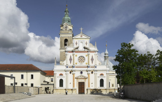 This photo shows the front view of a large church with an ornate white façade, decorative details, and a tall bell tower topped with a green dome. In front of the church is a wide open square under a bright sky with scattered clouds.
