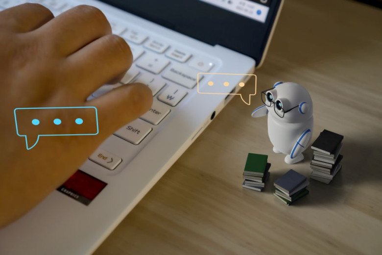 Hand typing on a laptop next to a small white robot surrounded by small stacks of books. Speech bubbles are floating above the keyboard and the robot indicating communication.