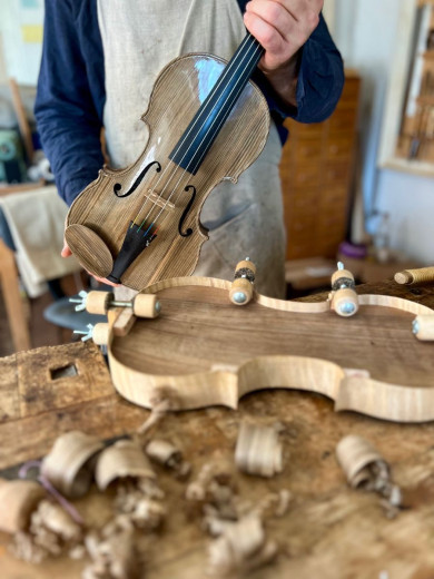 A person in an apron holds a hand-carved wooden violin. The workshop table displays wood shavings and a violin mold.
