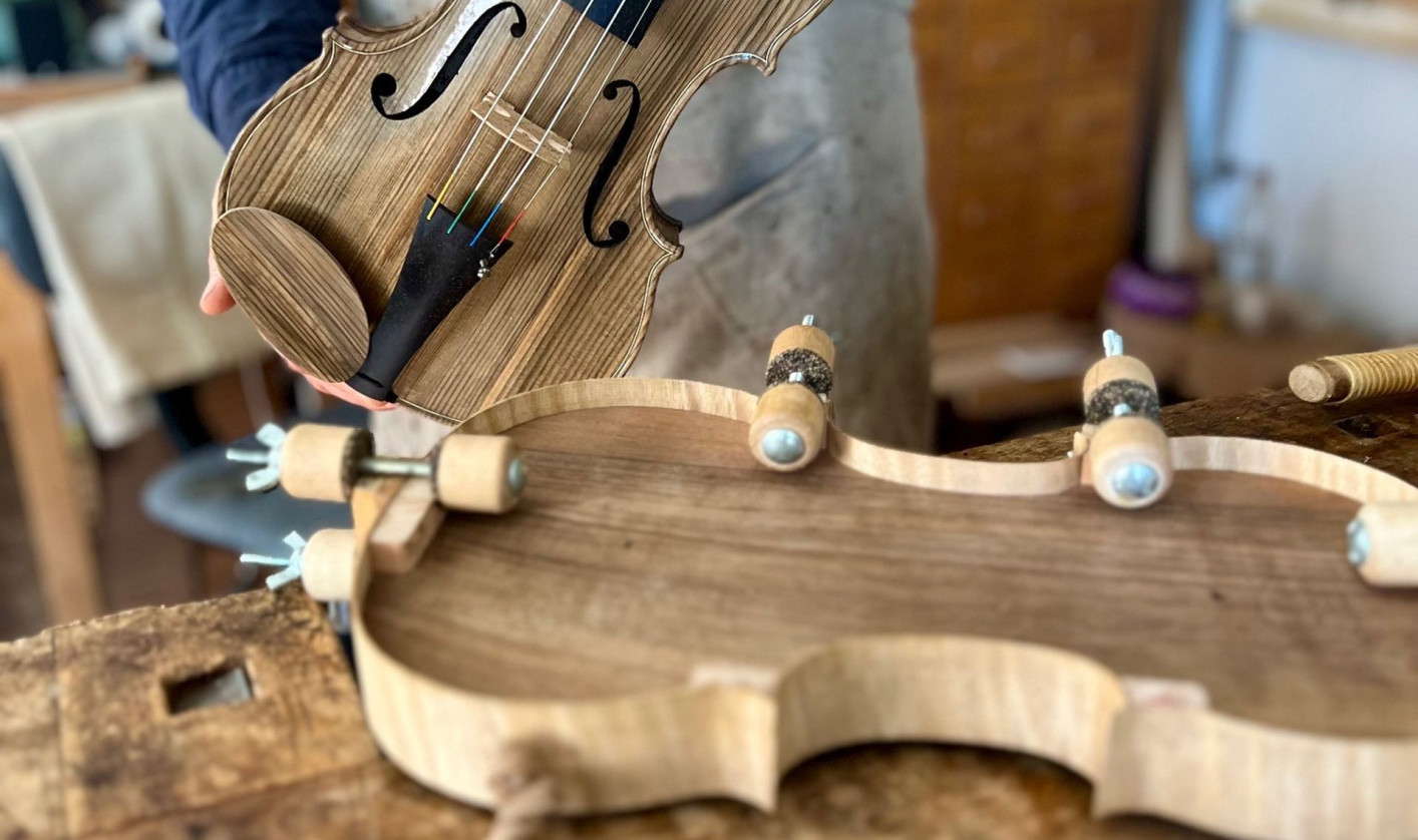 A person in an apron holds a hand-carved wooden violin. The workshop table displays wood shavings and a violin mold.
