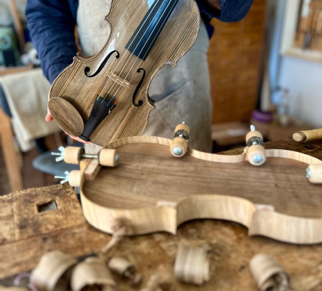 Oseba v predpasniku drži ročno izrezljano leseno violino. Na delovni mizi v delavnici so vidni lesni oblanci in kalup za violino.