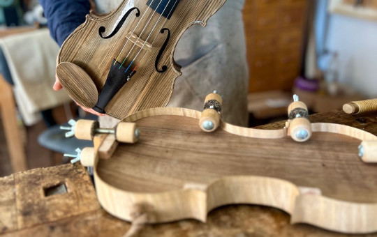 A person in an apron holds a hand-carved wooden violin. The workshop table displays wood shavings and a violin mold.