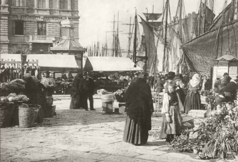 Historic black-and-white harbor market in Trieste with vendors, shoppers, produce baskets, and tall sailing ships lining the waterfront.