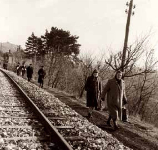 A group of women carrying milk containers walks beside a railway track, passing bare trees.
