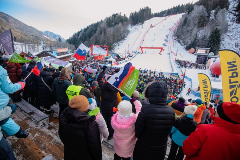 A vibrant crowd gathers at a snowy ski slope, waving Slovene and I feel Slovenia flags and dressed warmly.