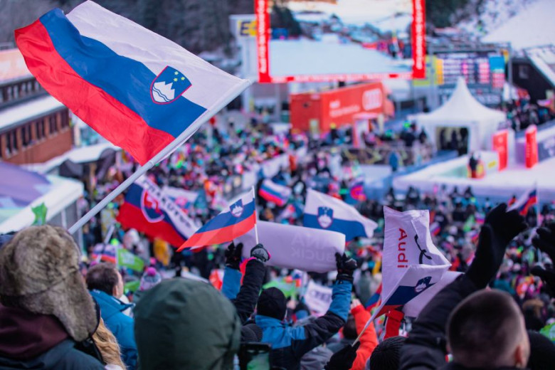 Crowd at a winter sports event, waving Slovenian flags with excitement. Snowy landscape, bright screens in the background.