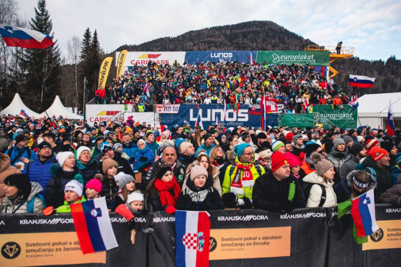A large, enthusiastic crowd in winter clothing gathers at the finish area of a ski race, surrounded by flags and banners, with a mountainous backdrop.