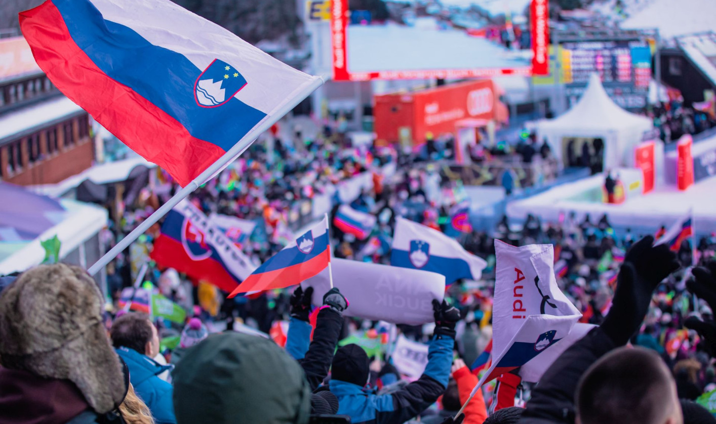 Crowd at a winter sports event, waving Slovenian flags with excitement. Snowy landscape in the background..