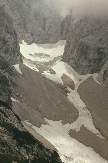 Photo shows a valley in the mountains, the peaks of the mountains are covered in mist, the bottom of the valley in partially covered in snow. 