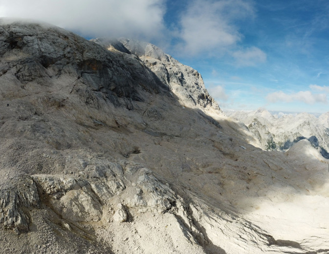A steep grey mountain slope with barely visible small patches of ice. The mountaintops are coveres in clouds with blue skies in the background.