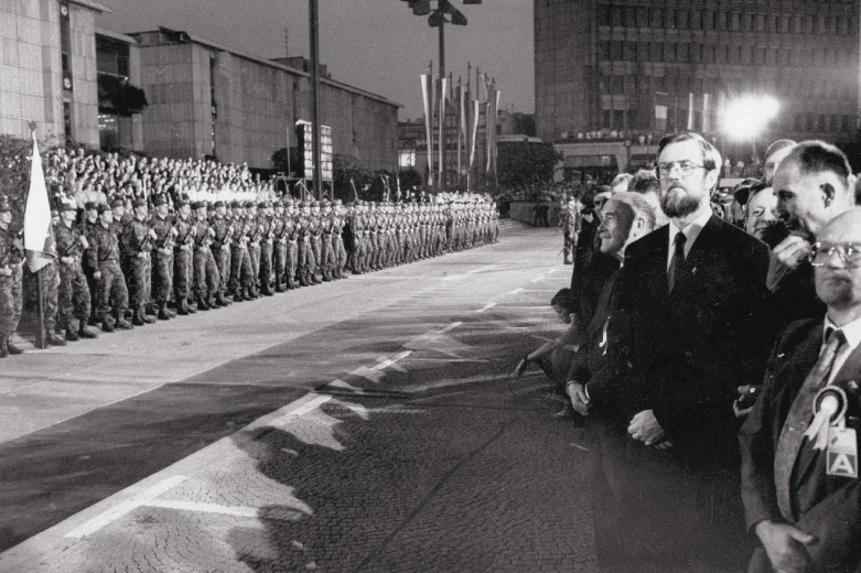 A black and white photo of a ceremony at night on the square in Ljubljana. On one side of the square are soldiers, lined up in multiple rows, while politicians and other people are standing onthe other side.