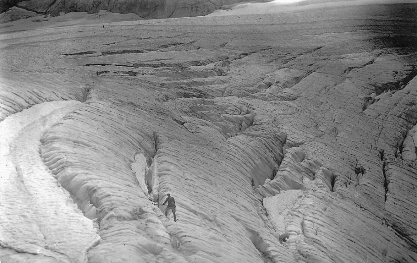A black and white photo shows a mountaineer walking across the heavily crevassed surface of the Triglav Glacier, with deep ice cracks and ridges stretching across the scene. 