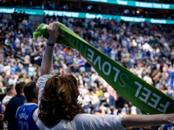 Ženska na tribunah, fotografirana od zadaj, v zraku drži zelen navijaški šal I Feel Slovenia.