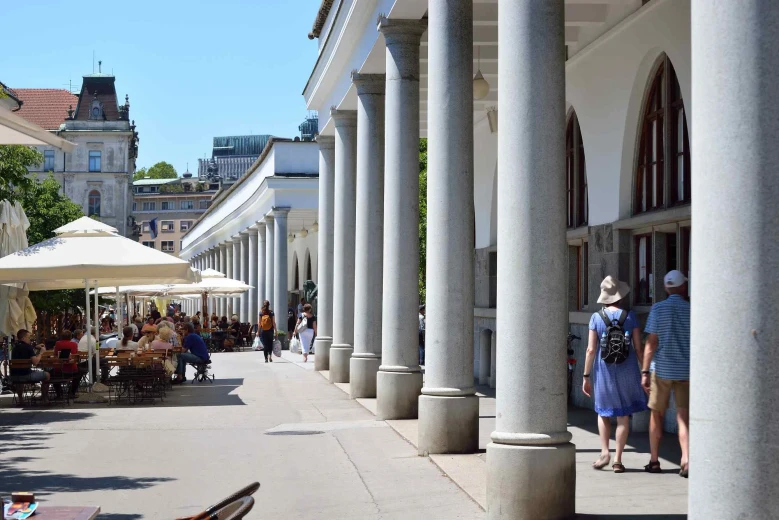 Ljubljana Market, capturing many white pillars or arcades. There are people sitting near at a bar outside on a sunny day.