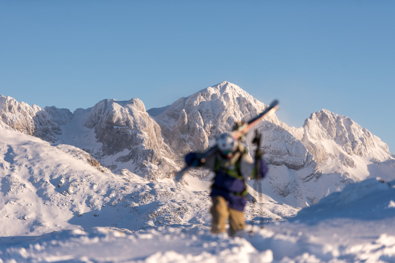 A downhill skier carries skis on his shoulders, with a mountain in the background.