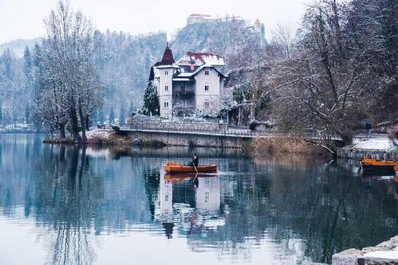 Blejsko jezero pozimi. V ozadju Blejski grad, v ospredju čoln na jezeru, ki ga vesla oseba na njem.