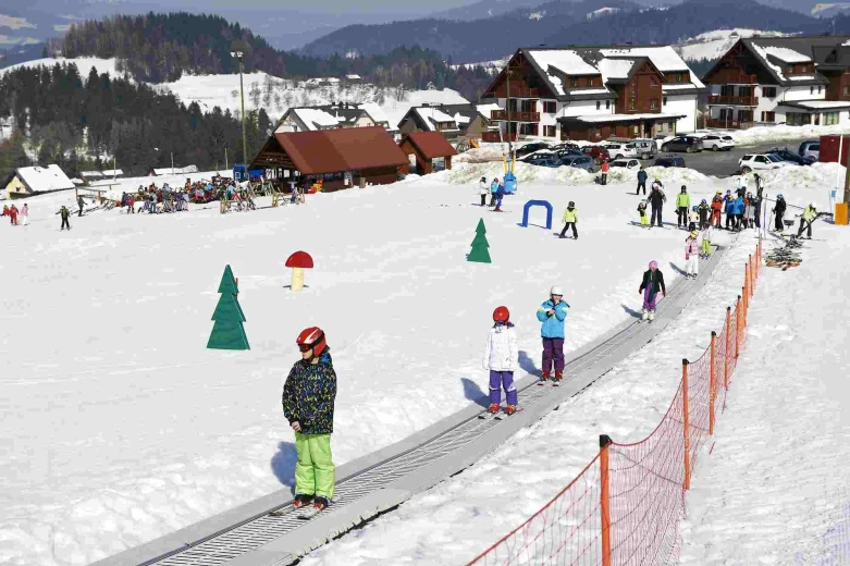 Otroci v smučarski opremi smučajo po ravni snežni progi ob rdeči ograji. V ozadju so koče in hoteli.