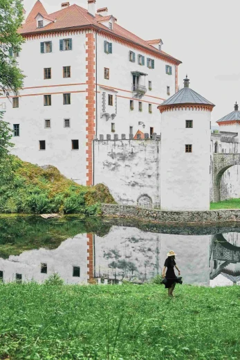 Snežnik Castle - a tall white castle with orange corners, standing behind a lake. A woman in a black dress is walking to the lake.