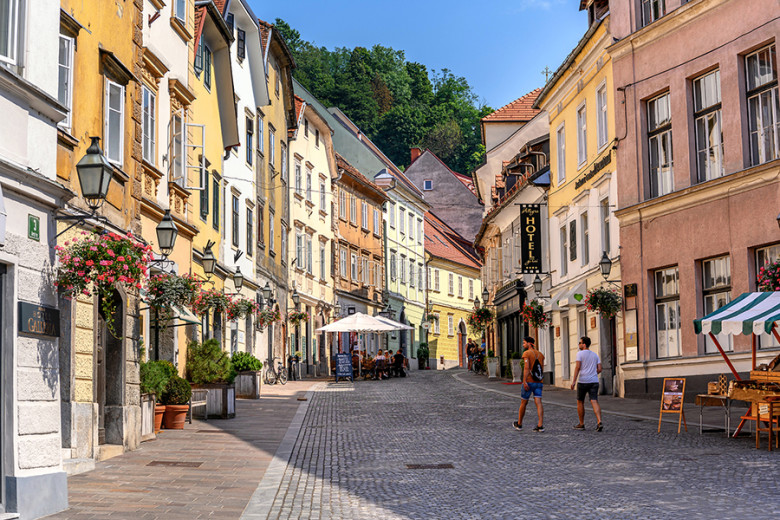 Tlakovana ulica v starem mestnem jedru Ljubljane s svetlo obarvanimi hišami, tesno druga ob drugi.