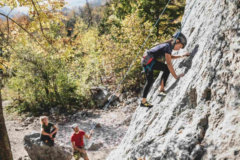 A little girl is climbing on a boulder, below is a coach holding the rope and another girl observing while sitting on a rock.