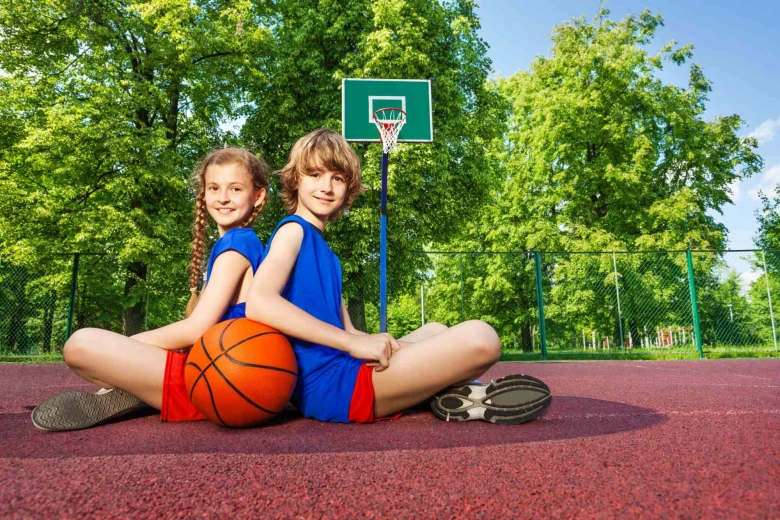 A boy and a girl are sitting on a basketball court outside, with a ball in front of them.