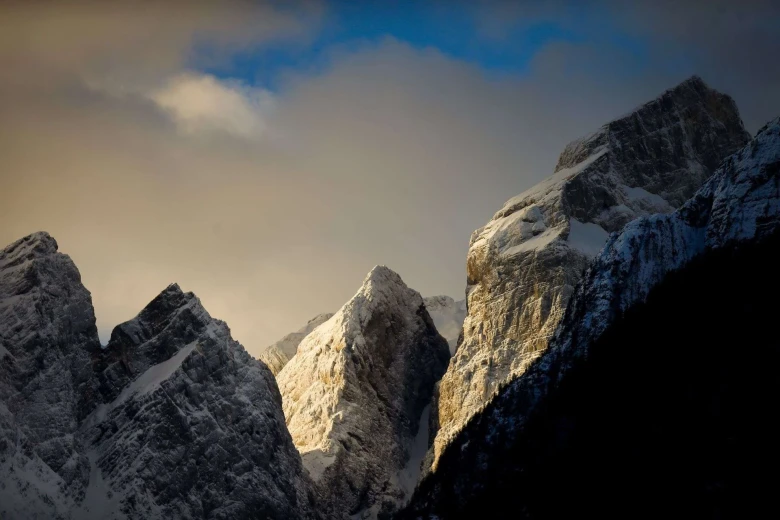 Panoramski pogled na zasnežene vrhove gora, pritisnjene ob oblačno nebo, in jutranje sonce, ki sije na eno stran gore na sredini.