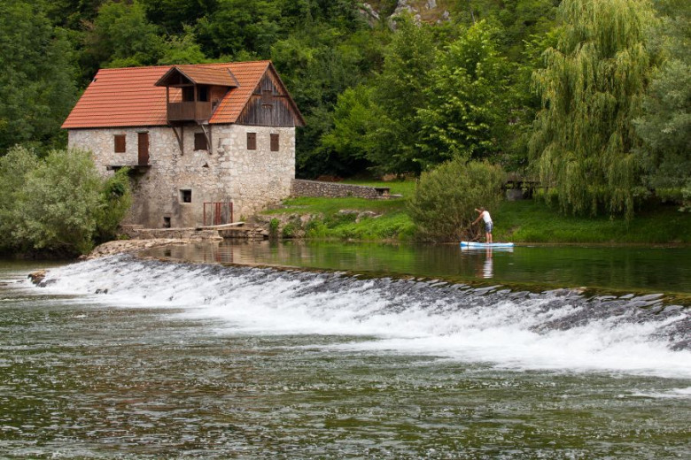 Na fotografiji je moški, ki supa po rekli Kolpi, pred njim pa so manjše brzice, v ozadju pa stoji stari mlin.